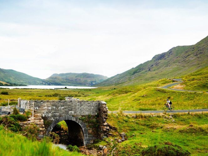 Cyclist riding toward a stone bridge flanked by green fields