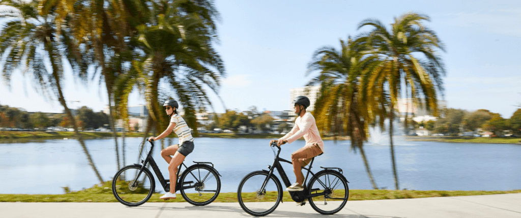 two people riding e-bikes on a paved, waterfront path with palm trees in the background