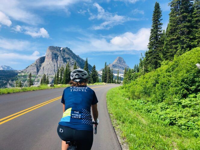 A single cyclists on a tree-lined road, riding toward mountains