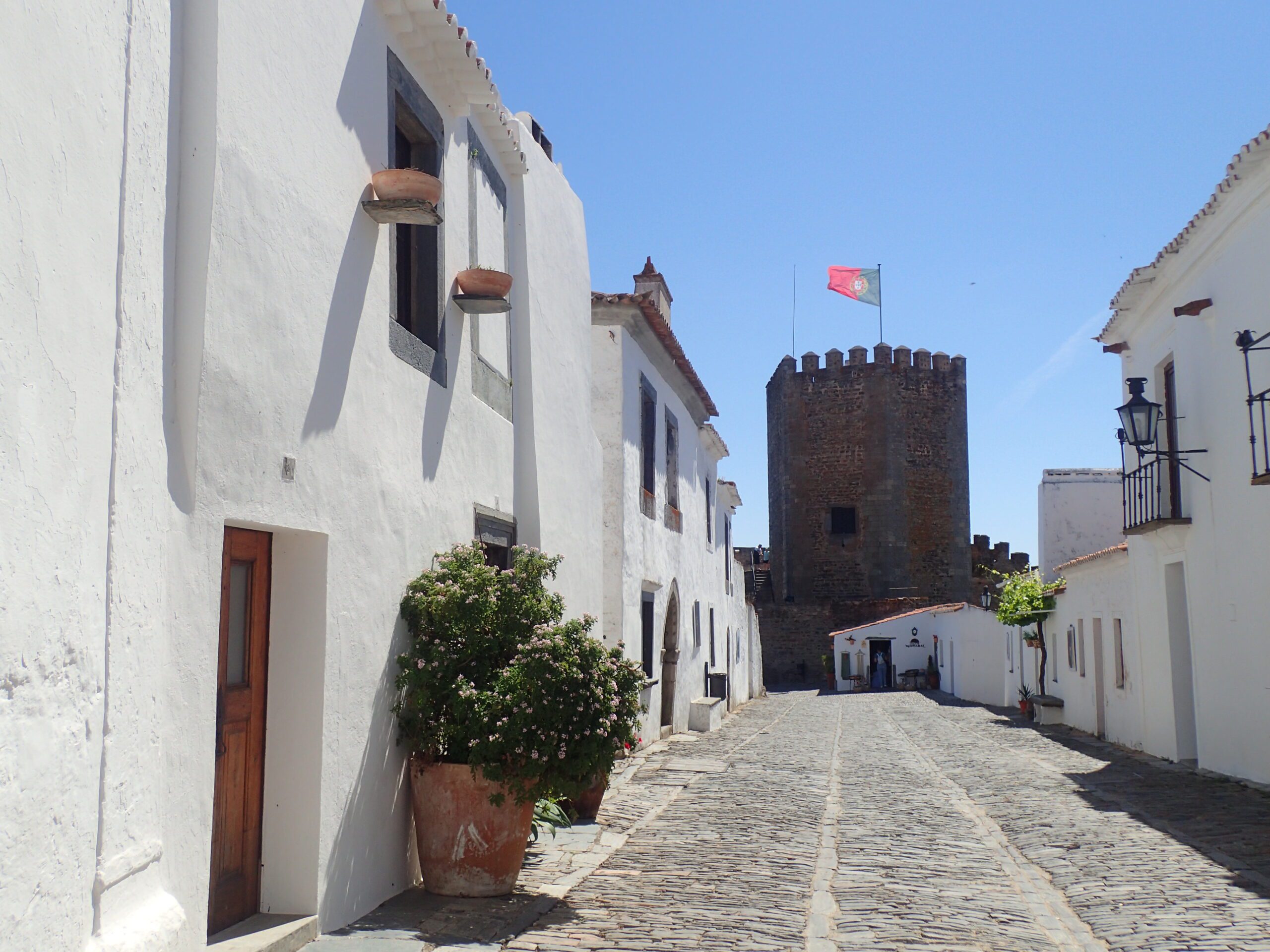 A quiet cobbled street in Monsaraz with white buildings and castle with Portuguese flag