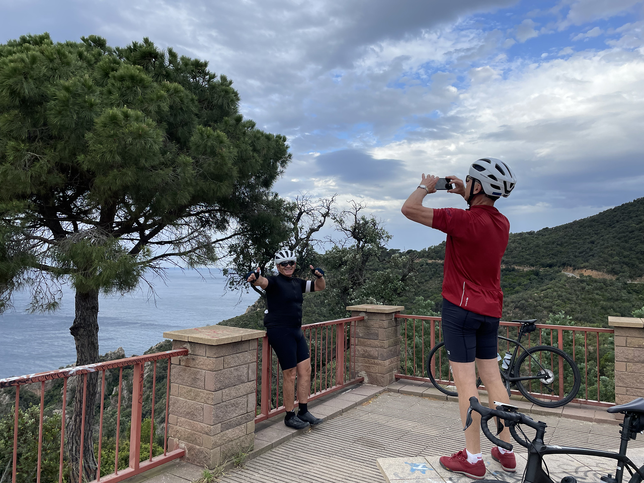 one cyclist taking a photo of another cyclist who is posing on a gate in Costa Brava with the sea in the background