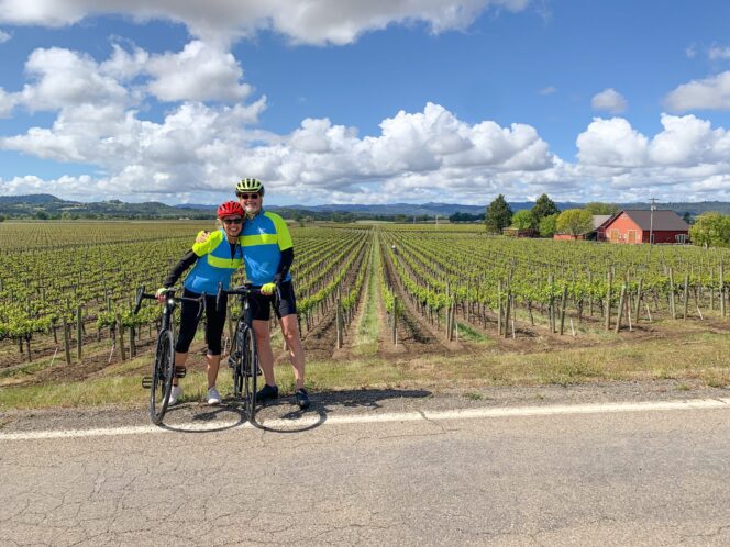 A couple pose with their bikes in front of rows of grape vines