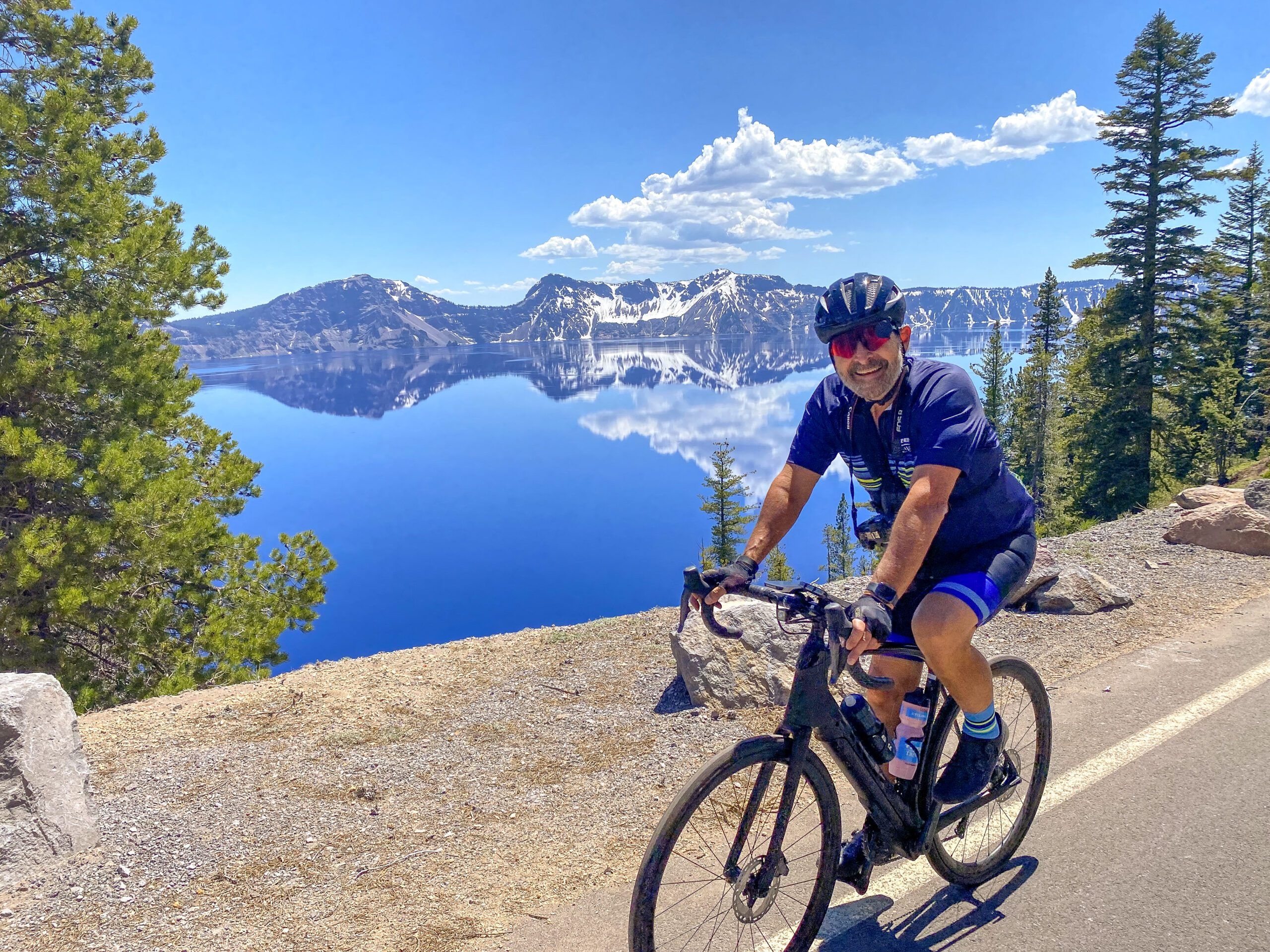 A cyclist rides on the road with Crater Lake and the volcano rim in the background.