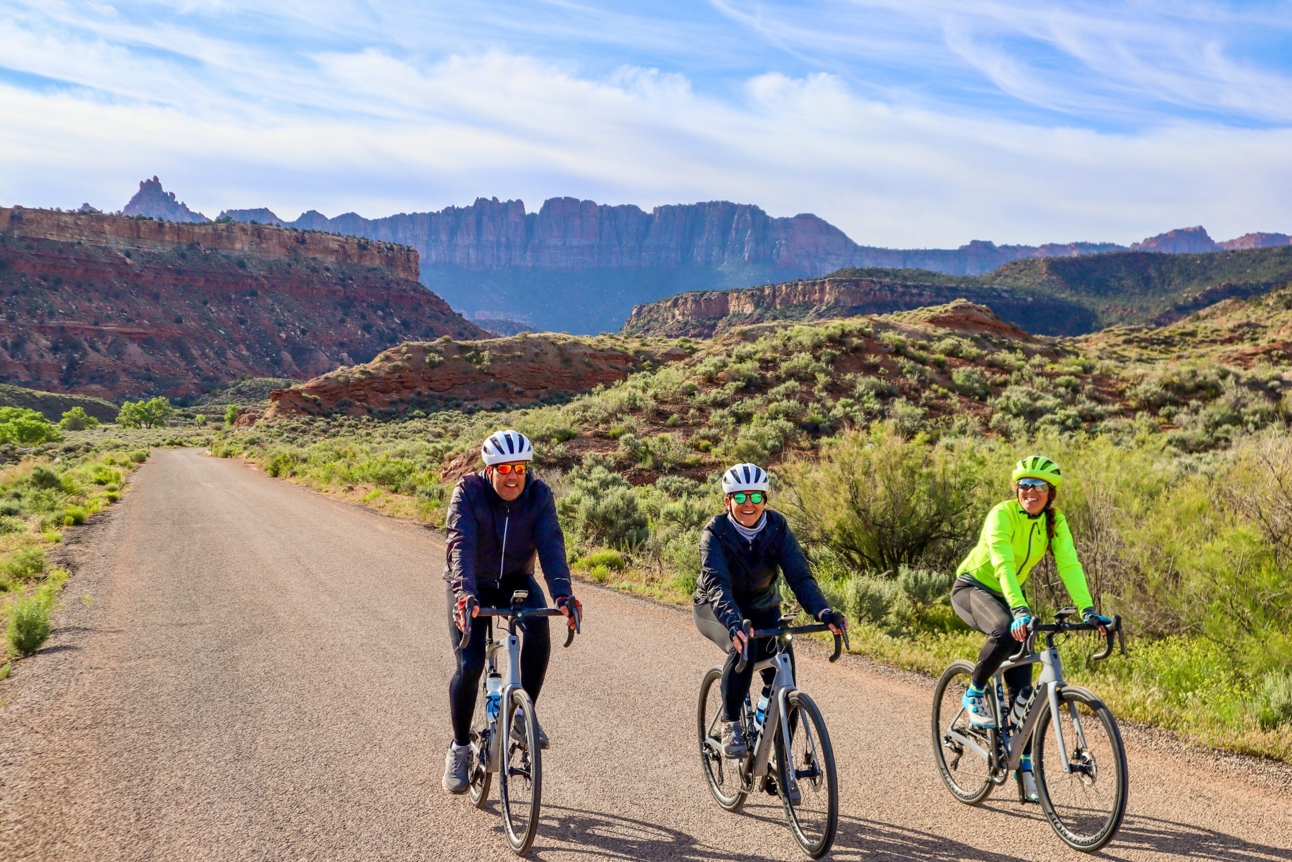 Three cyclists ride on a road with mesa rock formations in the background