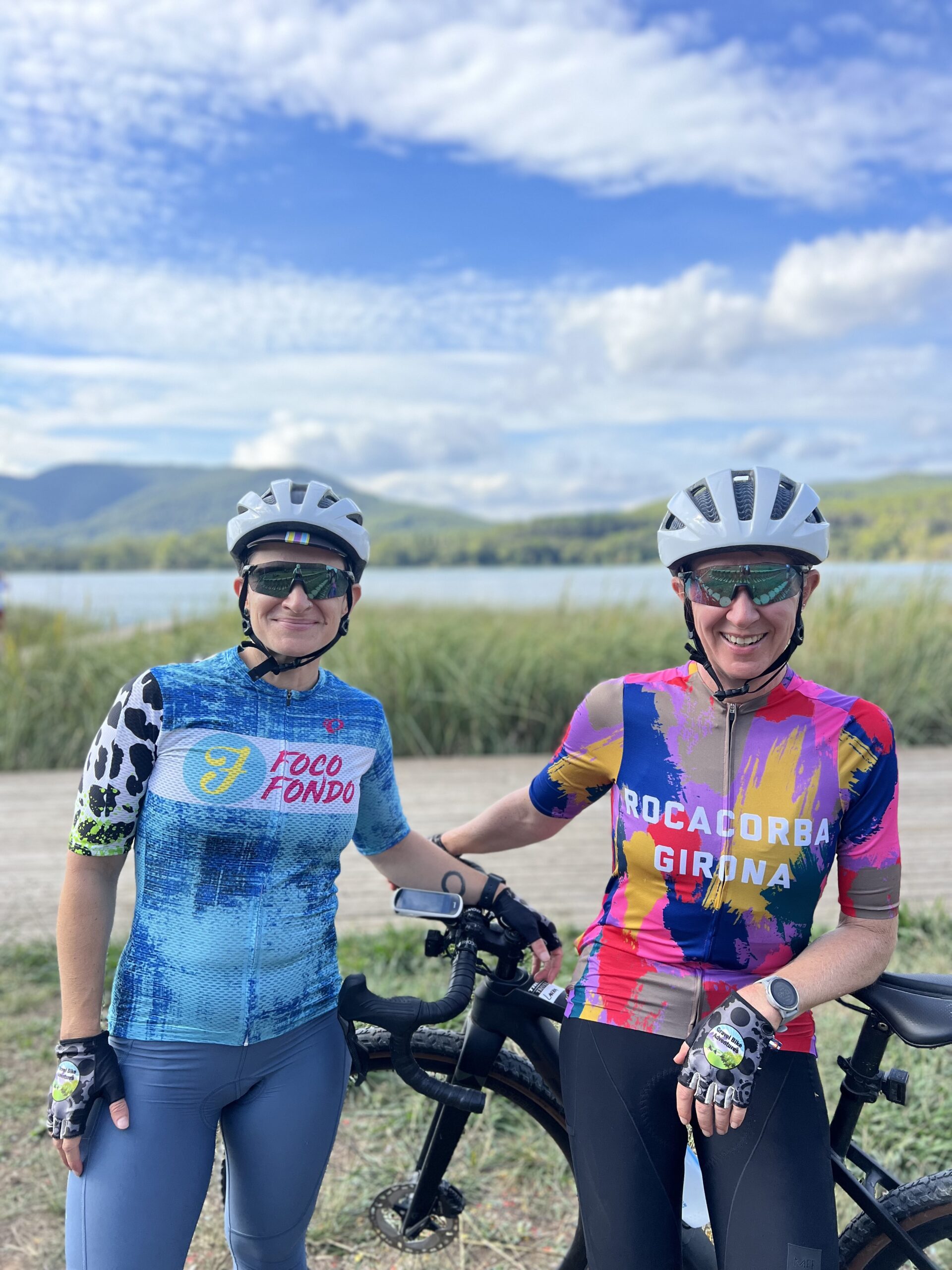 Two cyclists pose with their bikes with an out of focus lake and grasslands in the backgroun