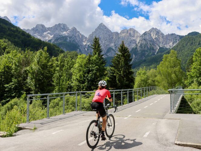 A cyclist rides toward a road bridge with the jagged Alps in the background