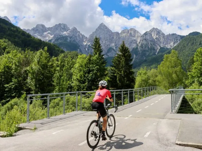 A cyclist rides toward a road bridge with the jagged Alps in the background