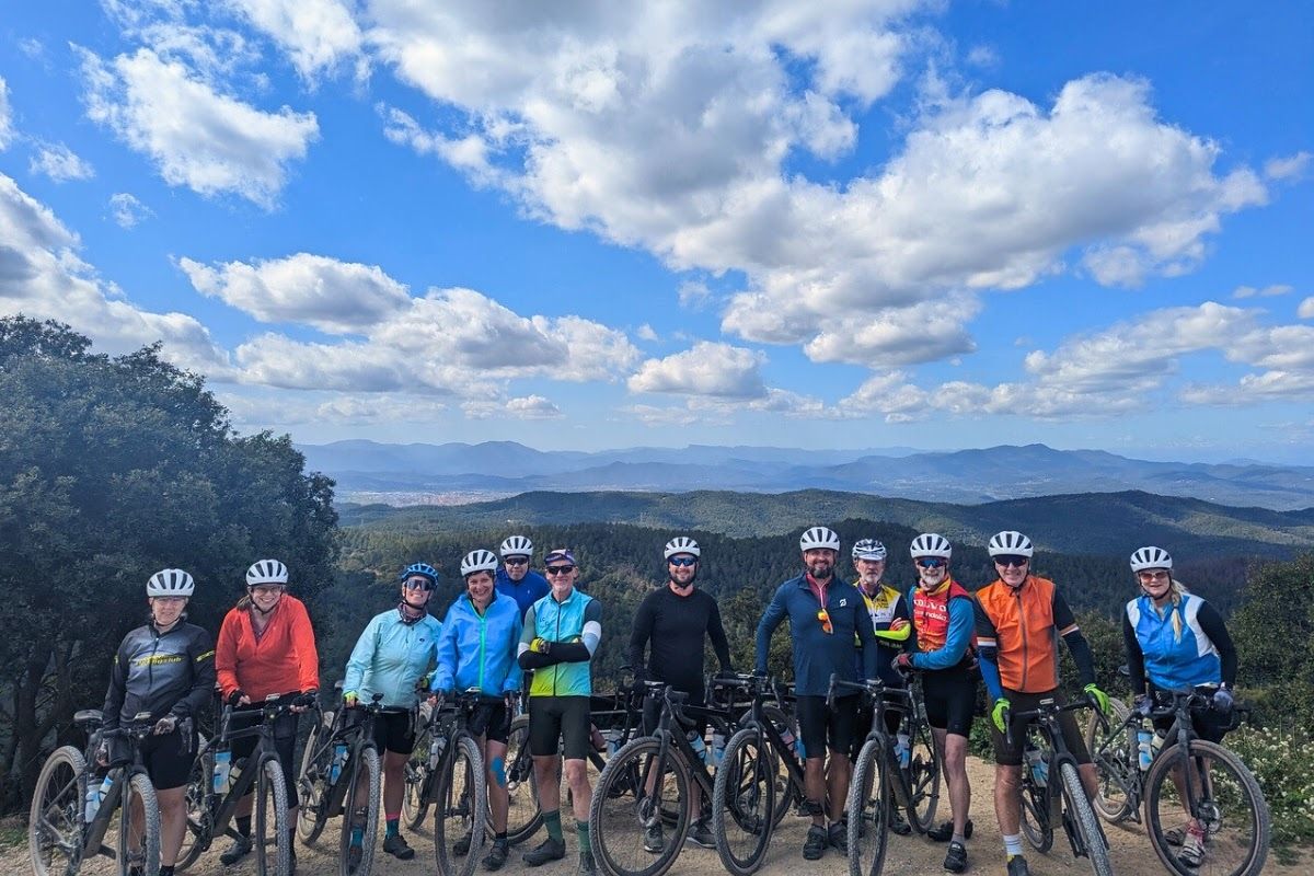 A group of cyclists pose in a line in front of tree-covered hills