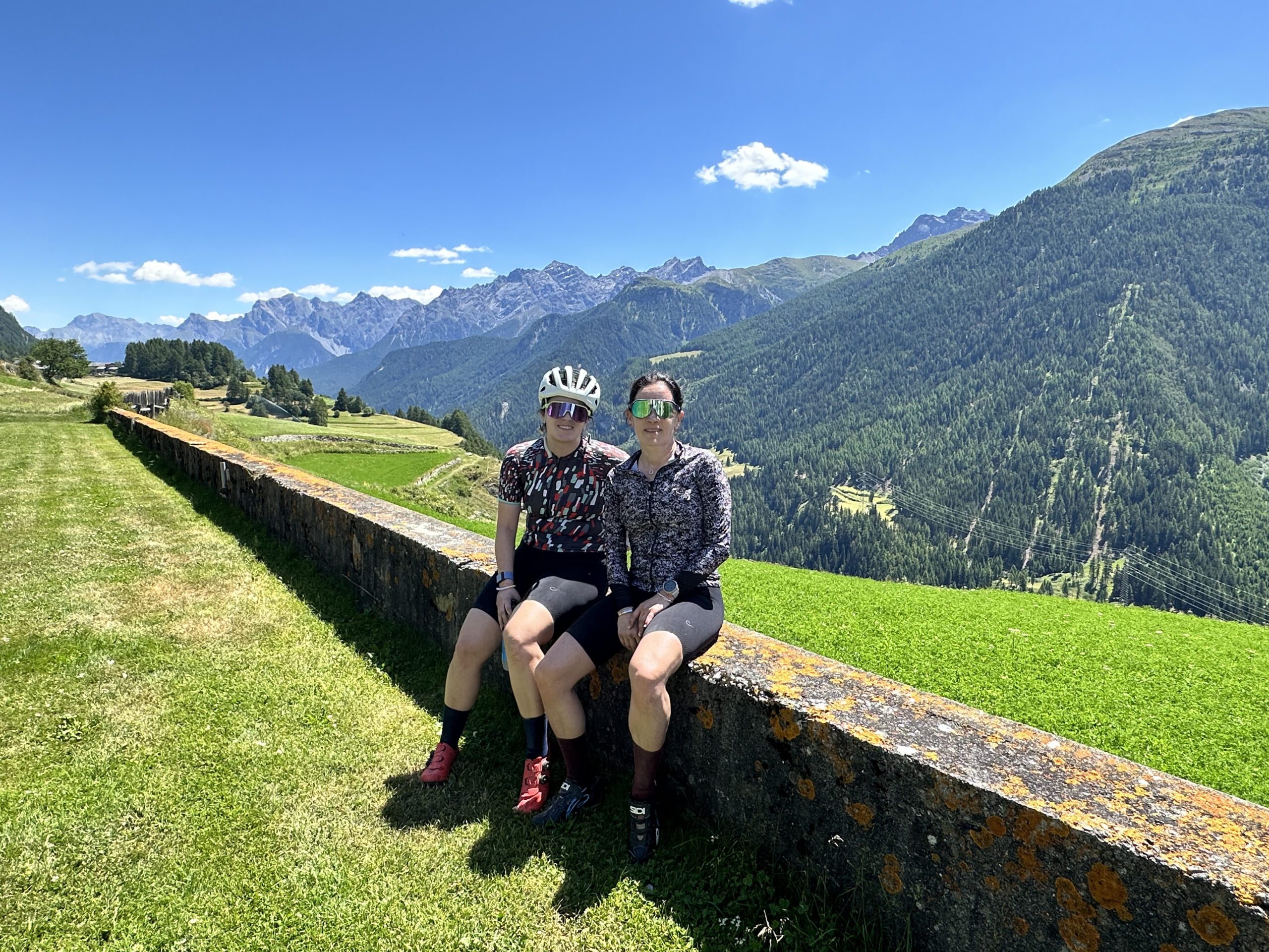 Two cyclists sit on a low stone wall with the Alps in the background