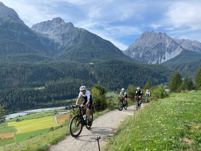 Four cyclists on a gravel track above a river valley with mountains