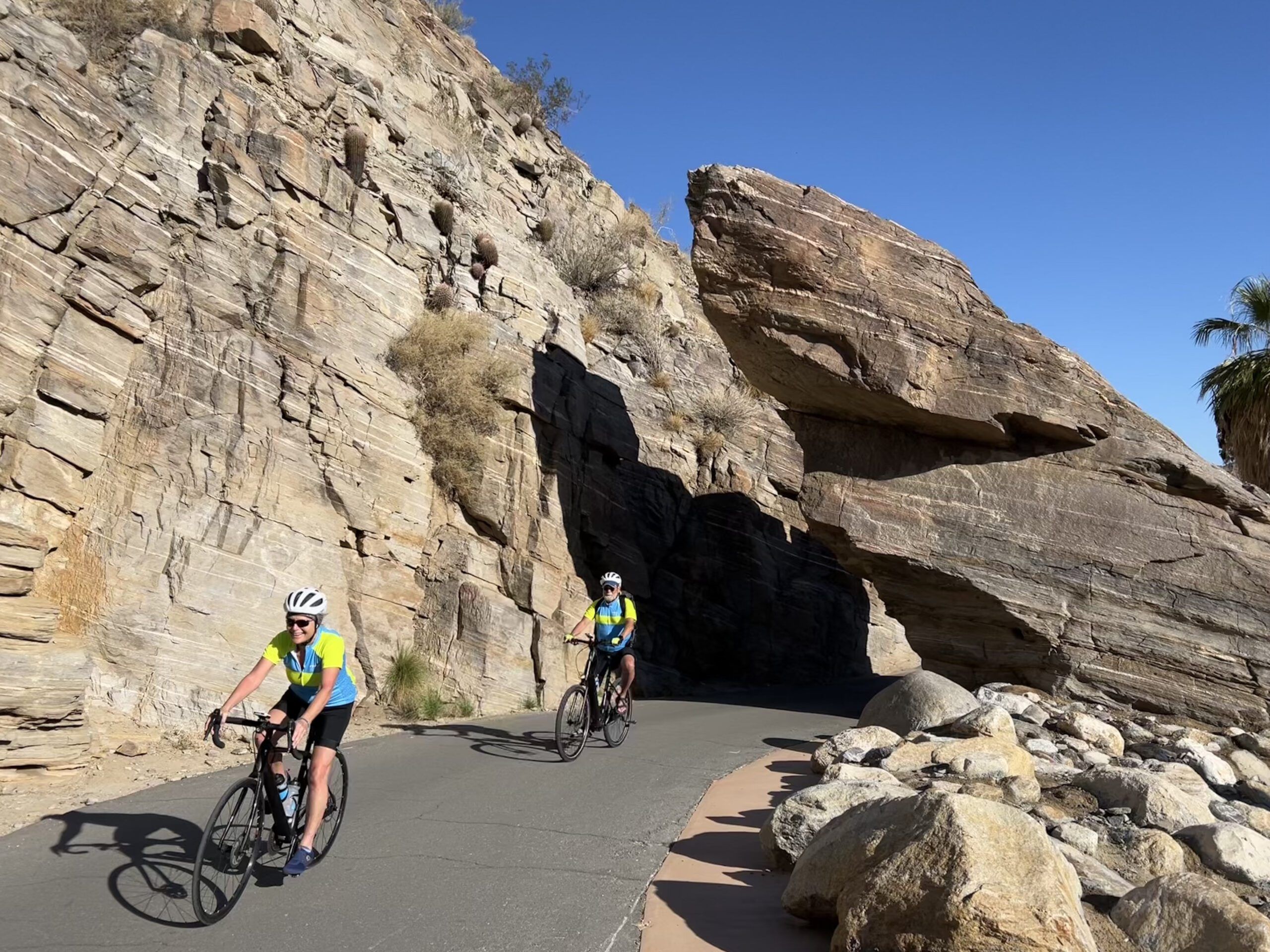 Two cyclists ride in front of a rock wall under blue skies