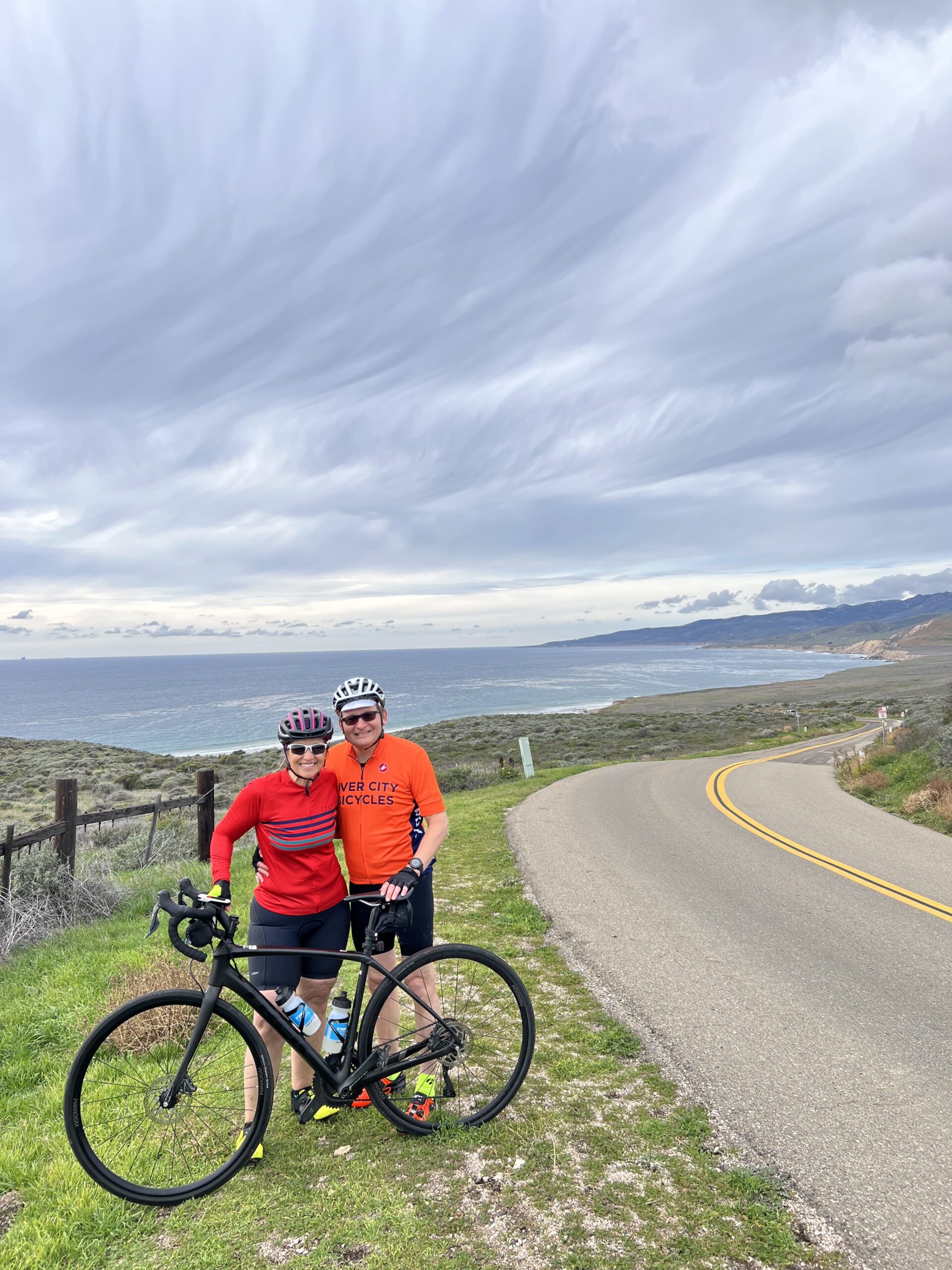 Two cyclists pose by the side of the road overlooking the ocean