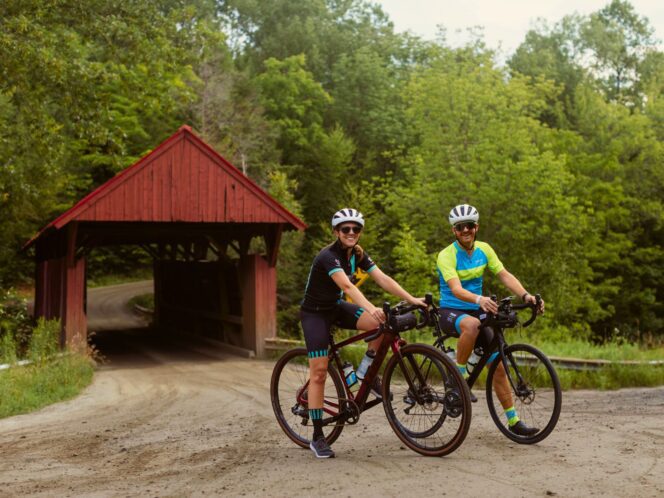 Two cyclists on a gravel road with a covered bridge and forest in the background