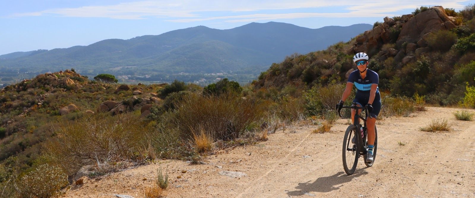 A female cyclist rides a hard-packed gravel road with mountains in the background