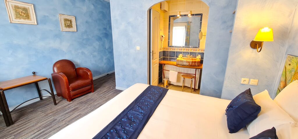 Double bedroom with tiled bathroom and armchair at Hotel du Mas