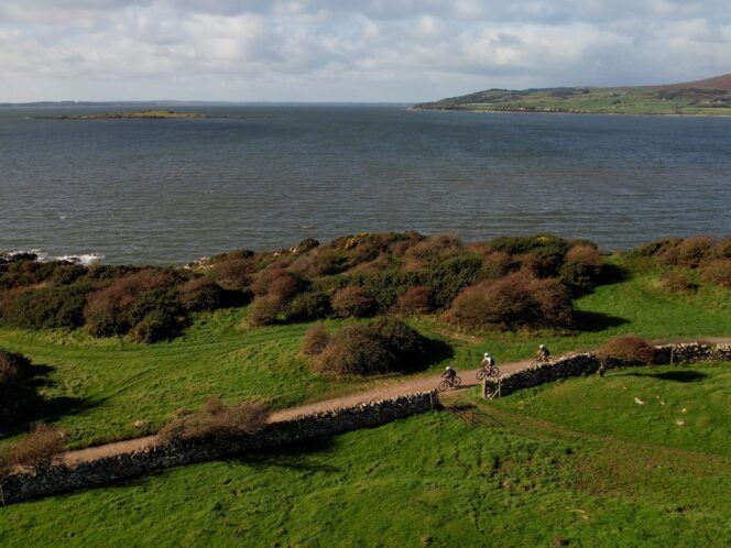 Aerial view of three cyclists on a gravel path with stone walls and a lake